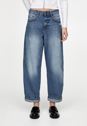 Kvinde iført opsmøgede, løstsiddende blå jeans med let slid samt sorte loafers, kombineret med en hvid cropped ribbet top.