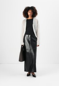 Lett og gjennomsiktig hvit cardigan over en svart ribbestrikket topp; svart skinn midi-skjørt; holder en brun tote bag; svarte flate sko.