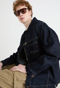 Mørk denimjakke med gullsøm, med to brystlommer. Modellen har på seg brune solbriller og beige cargo-bukser.