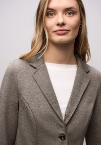 Szary, teksturowany marynarka z klapami i pojedynczym zapięciem na guzik, noszona na białym topie. Zbliżenie na szczegóły ubrania.