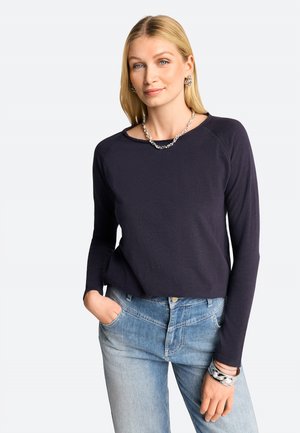 Navyblaues Langarmoberteil aus weichem Stoff mit lässiger Passform, Rundhalskragen und Raglanärmeln, kombiniert mit hellblauen Jeans und silbernem Schmuck.