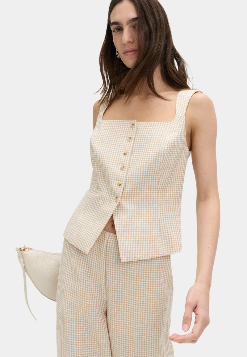 Woman wearing beige and white checkered sleeveless top with buttons and matching wide-leg pants, holding a small cream handbag.