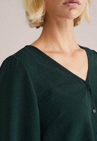 Top verde a scollo a V realizzato in un tessuto tessurizzato, dotato di maniche corte a palloncino e due bottoni neri lungo la patta.