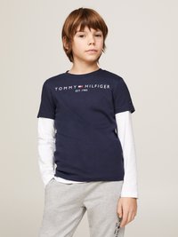 Navy Baumwoll-T-Shirt mit dem Logo "TOMMY HILFIGER EST. 1985", darüber ein weißes Langarmhemd, kombiniert mit grauen Sweatpants.