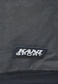 Čierna textúrovaná taška z látky s obdĺžnikovou čiernou gumovou nášivkou s bielym textom "KANI SPORT" a kontrastným lemom.