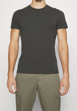 Mand iført en tætsiddende sort T-shirt med rund hals og olivengrønne cargo-bukser stående mod en ensfarvet baggrund.