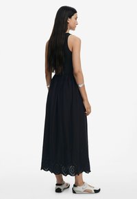 Vestido maxi negro con un cuerpo ajustado y una falda fluida, con detalles de dobladillo cortados con láser intrincados. Llevado con calzado deportivo de color claro.
