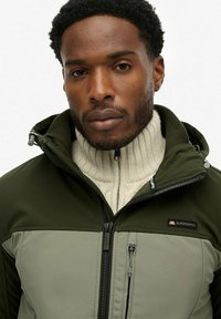 Olijfgroene en lichtgrijze jas van zacht, weerbestendig materiaal. Hoge kraag, ritssluiting en een geribbelde crèmekleurige trui eronder.