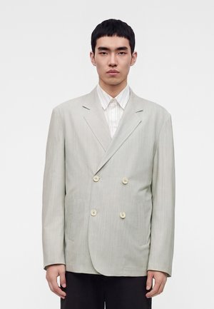 Jeune homme aux cheveux noirs courts portant un blazer gris clair double boutonnage sur une chemise blanche à col, sur un fond uni.