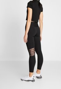 Leggings neri con pannelli in rete e un design di logo ripetuto, abbinati a un top corto nero e sneakers bianche con una suola a motivi.