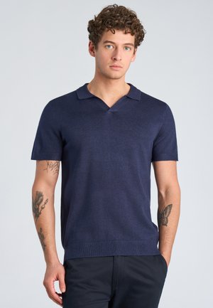 Jeune homme aux cheveux bouclés portant une chemise à col bleu marine à manches courtes, mains dans les poches, montrant des tatouages sur les deux avant-bras.