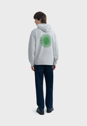 Jonge man gekeerd met zijn rug naar voren, draagt een lichtgrijze hoodie met een groene zonnestraal ontwerp en donkerblauwe spijkerbroek.
