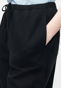 Κοντινό πλάνο ενός ατόμου που φοράει μαύρο παντελόνι με κορδόνι και ελαστική μέση, με το χέρι εν μέρει μέσα στην πλαϊνή τσέπη.