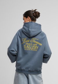 Plava hoodica s velikim žutim izvezenim tekstom na leđima "Lost Treasures Pequs Art Gallery Museum, Mykonos." Mekani materijal, opušteni kroj.
