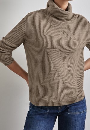 Personne portant un pull à col roulé côtelé beige avec un motif géométrique texturé, associé à un jean en denim bleu.