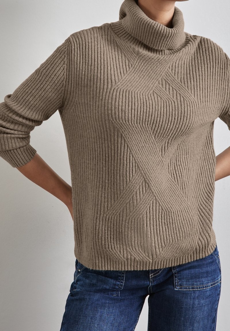 Personne portant un pull à col roulé côtelé beige avec un motif géométrique texturé, associé à un jean en denim bleu.