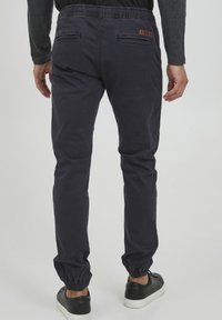 Marineblå joggingbukser med elastiske manchetter og linning, med baglommer og en lille brun læderdetalje. Glat tekstur, afslappet design.