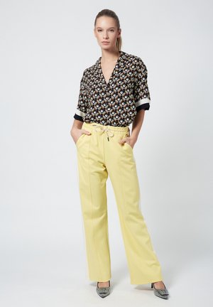 Black and white patterned blouse with a collar and rolled sleeves, paired with high-waisted yellow trousers with a drawstring, worn with heeled shoes.