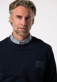 Sweatshirt bleu marine avec un col rond et un patch en caoutchouc portant le logo BOSTON PARK. Porté par-dessus une chemise à carreaux bleue et jaune avec un col.