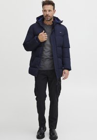 Marineblå puffjakke med hette, quiltet design, med blå fôr. Brukt over en mørkegrå skjorte og svarte cargo-bukser, sammen med svarte støvler.