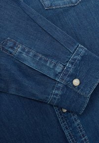 Puño de camisa de mezclilla azul de mangas largas con botón a presión blanco y detalle de doble costura visible.