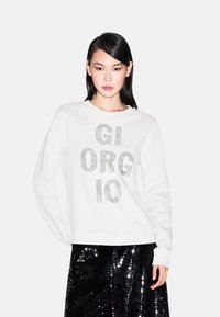 Sudadera blanca con gran texto de piedras de strass plateadas "GIORGIO" en el centro, combinada con una brillante falda negra de lentejuelas.