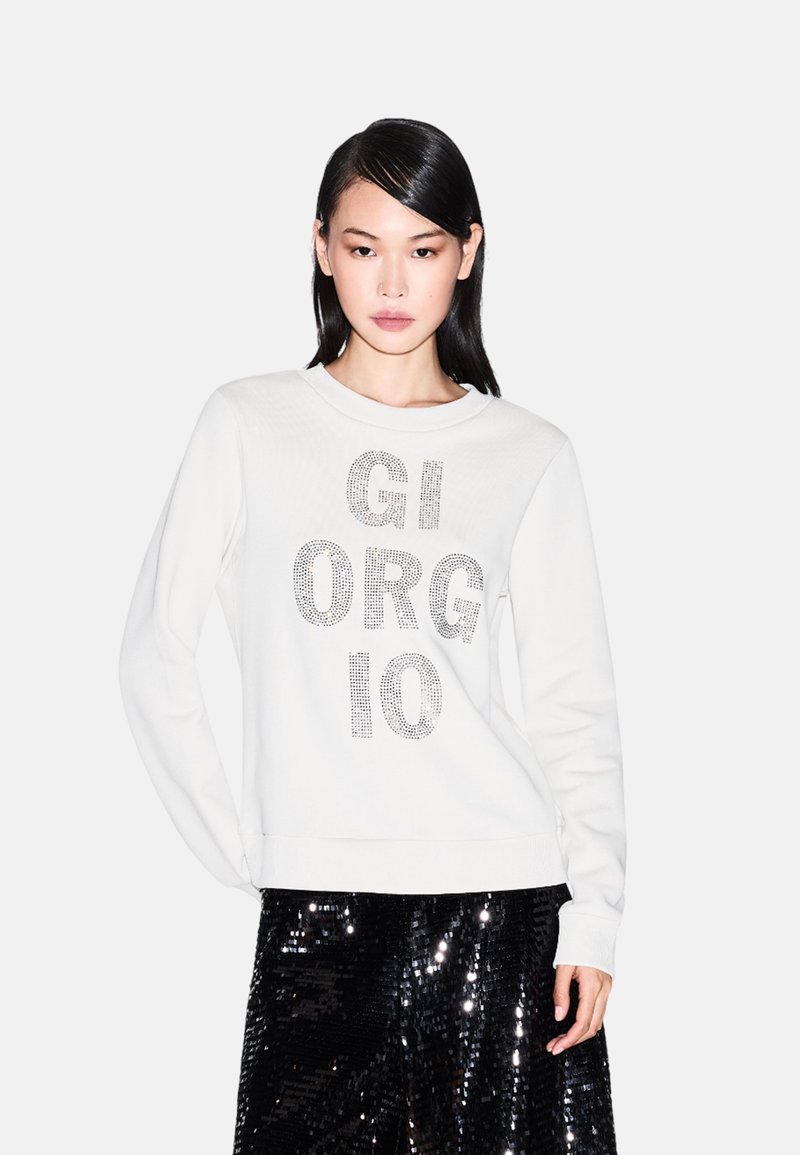 Sudadera blanca con gran texto de piedras de strass plateadas "GIORGIO" en el centro, combinada con una brillante falda negra de lentejuelas.