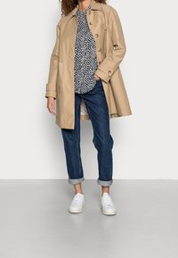 Nainen yllään beige trenssitakki, mustavalkoinen polkadottipaita, käärityt siniset farkut ja valkoiset tennarit, seisoo yksiväristä taustaa vasten.