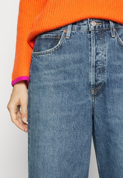 Blue denim jeans with a straight cut, featuring a metal button fly, five pockets, and a slightly faded texture. Bright orange knitted sweater.