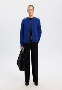 Cardigan de punto azul con cuello redondo, botones en el frente y mangas largas. Combinado con una blusa negra, pantalones anchos negros y un gran bolso negro.