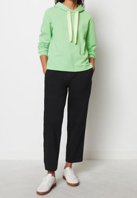 Vaaleanvihreä hupullinen huppari, jossa kiristysnyöri, yhdistettynä mustiin housuihin. Asun täydentävät valkoiset lenkkarit ruskeilla pohjilla. Pehmeä ja tasainen pinta.