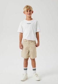 T-shirt blanc avec le texte "Édition studio" en noir, shorts beiges et baskets blanches. Chaussettes rayées noir et blanc. Style décontracté.