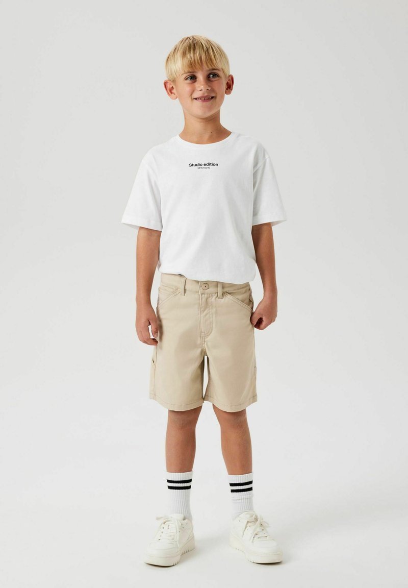 T-shirt blanc avec le texte "Édition studio" en noir, shorts beiges et baskets blanches. Chaussettes rayées noir et blanc. Style décontracté.