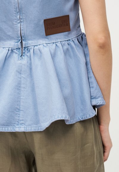 Vue de dos d'une personne portant un top en denim bleu clair à volants avec une étiquette marron « Baum und Pferdgarten » et un pantalon vert olive.
