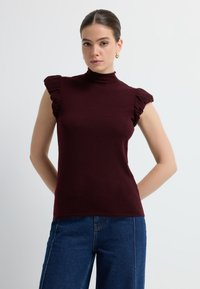 Top sin mangas burdeos con cuello alto y detalle de hombros con volantes, hecho de una tela suave. Combinado con unos vaqueros de pierna ancha azul.