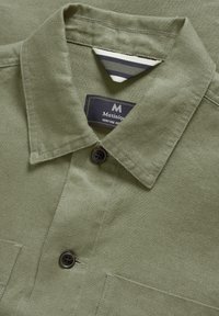 Camisa verde oliva con cuello, que presenta una etiqueta tejida y un cierre de un botón. Tejido texturizado con detalles de costura visibles.