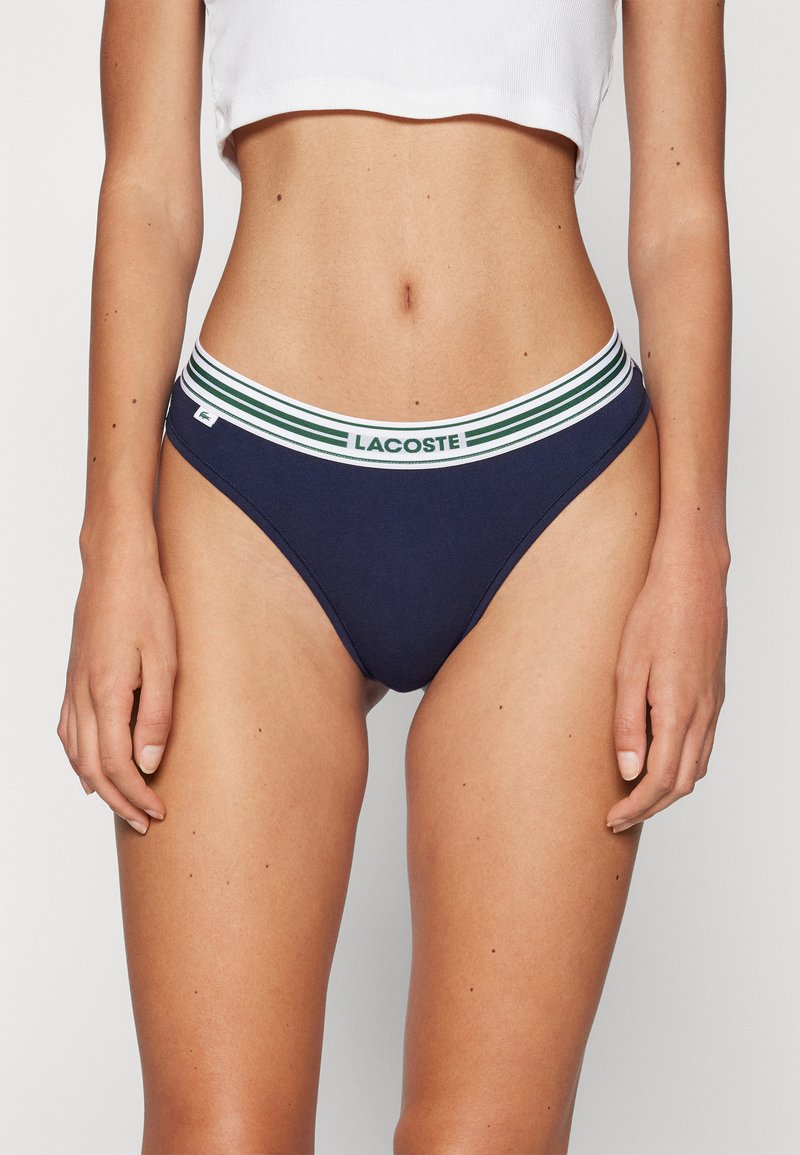 Bikini puuvillasta tummansinisellä värillä, jossa on valkoinen ja vihreä raidallinen vyötärö. Edessä on logolappu ja se on sileä, hyvin istuva malli.
