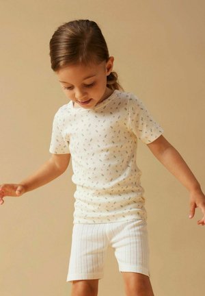 Nuori tyttö vaaleanruskeilla hiuksilla, valkoinen lyhythihainen paita, jossa pieni kukkakuosi, ja valkoiset teksturoidut shortsit, katse alaspäin.