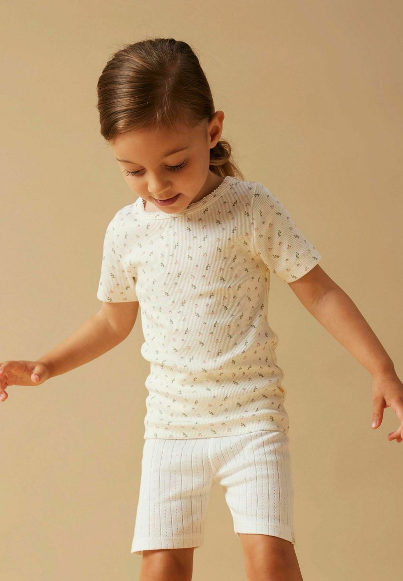 Junges Mädchen mit hellbraunen Haaren, das ein weißes kurzärmeliges Hemd mit kleinem Blumenmuster und weiße strukturierte Shorts trägt und nach unten schaut.