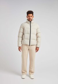 Lys grå pufferjakke med høj krave, udstyret med en central sort lynlås og quiltet design. Samlet med beige bukser og hvide sneakers.