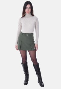 Maglione beige a collo alto, design aderente. Gonna plissettata verde scuro con cintura e fibbia argento. Stivali neri al ginocchio con finitura liscia.
