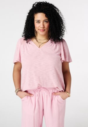 Femme aux cheveux bouclés noirs portant un chemisier rose à col en V et un pantalon rose, les mains dans les poches, accessoirisée avec des bijoux en or.