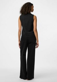 Juoda be rankovių palaidinė, prigludusio fasono su aukštu apykakle, derinama su juodomis plačiomis džinsinėmis kelnėmis, nešiotomis aukšta juosmeniu, matoma iš nugaros.