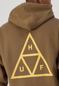 Brauner Kapuzenpulli mit einem großen gelben dreieckigen Grafik, die die Buchstaben "H", "U" und "F" enthält. Weicher Stoff, normale Passform.
