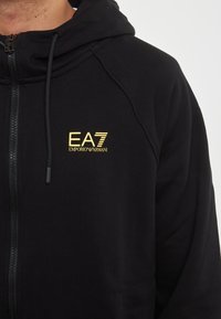 Felpa con zip nera in tessuto morbido, con il logo dorato EA7 Emporio Armani sul lato sinistro del petto e cappuccio con coulisse.