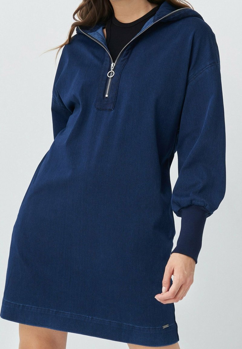 Femme portant une robe à capuche bleu marine ample avec une fermeture éclair argentée à l'avant et des manches longues avec des poignets ajustés.