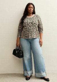 Beige en groene trui met panterprint, gecombineerd met lichtblauwe wijde jeans met opgerolde zoom. Model houdt een kleine zwarte handtas vast.