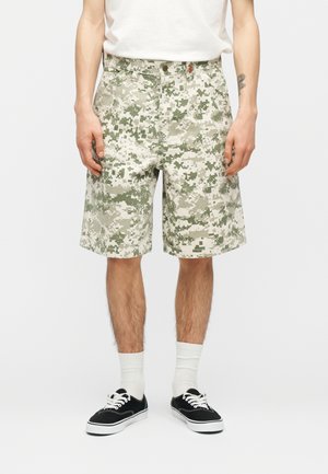 Homme portant un short camouflage numérique vert clair, des chaussettes blanches et des baskets noires, debout devant un fond uni.