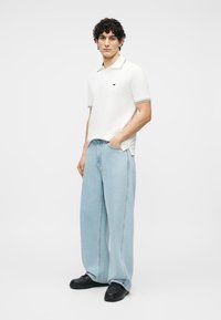 Νέος άνδρας που φοράει λευκό πόλο πουκάμισο, ανοιχτό μπλε τζιν με φαρδιά μπατζάκια και μαύρα παπούτσια, στέκεται με το ένα χέρι στην τσέπη μπροστά σε απλό φόντο.