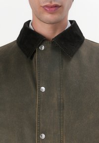 Olive green outerwear jacket with a corduroy collar, silver snap buttons, and a worn texture. Features a straight design with visible stitching.